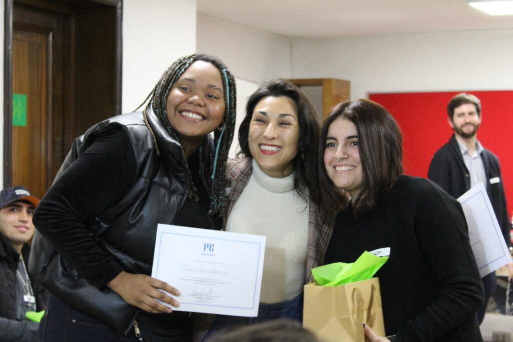 Trinidad Luengo, Presidenta de Fundación Proyecto B, entrega diploma de egreso con el regalo respectivo a Maira, beneficiaria de residencias de nuestra fundación, junto a Carolaine Aguila, asesora laboral. Las tres posan a la cámara sonriendo.  